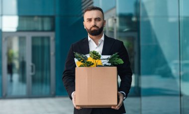 Dışarıda takım elbiseli üzgün bir işadamının bir kutu eşyayla dikilmiş portresi. Erkek ofis çalışanı işini kaybetti.