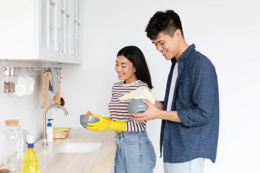 Asyalı adam, neşeli kız arkadaşının mutfakta bulaşık yıkamasına yardım ediyor.