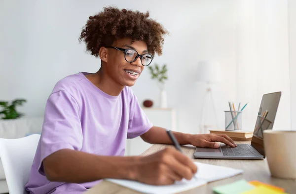 Conceito de ensino em casa. inteligente preto teen cara usando laptop na mesa, escrevendo no notebook durante on-line lição dentro de casa — Fotografia de Stock