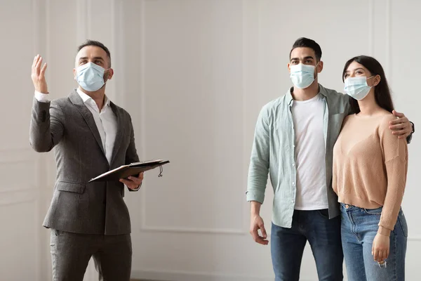 Агент по недвижимости в медицинской маске показывает покупателям новую квартиру — стоковое фото