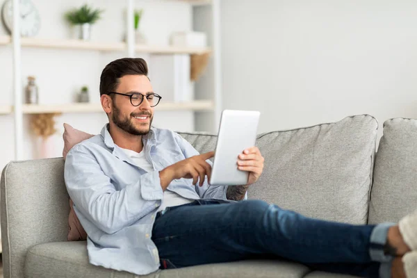 Koncepcja technologii. Uśmiechnięty tysiącletni mężczyzna korzystający z tabletu cyfrowego spoczywającego na kanapie w swoim nowoczesnym mieszkaniu, wolna przestrzeń — Zdjęcie stockowe