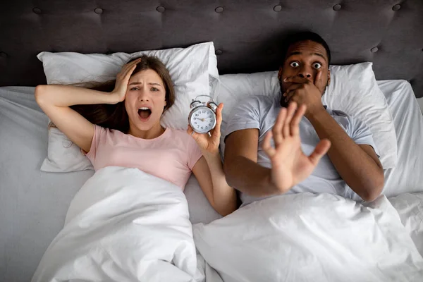 Visão superior do casal interracial estressado com despertador gritando de terror, atrasando-se para o trabalho, deitado na cama, vista superior — Fotografia de Stock