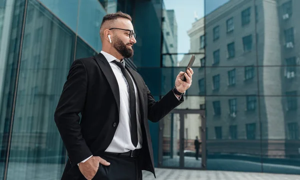 Boldog üzletember öltönyben beszél okostelefonon vezeték nélküli fülhallgatóval áll üveg irodaház mellett a szabadban — Stock Fotó