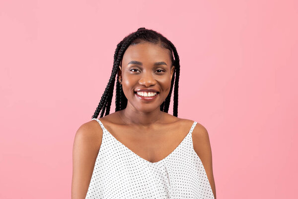 Portrait of happy black lady with afro bunches posing in summer dress, looking at camera on pink studio background