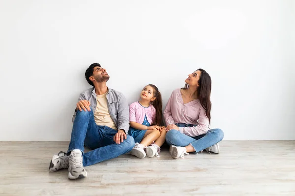 Pensando em design de interiores. família árabe feliz sentado no chão inclinado na parede branca e olhando para o espaço vazio — Fotografia de Stock