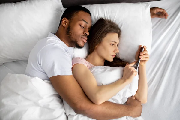 흑인 남편의 품에 안긴 채 침대에 누워 있는 젊은 여자가 자기 애인에게 문자를 보내기 위해 핸드폰을 사용하고 있습니다. — 스톡 사진