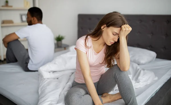 Interrassische Eheleute, die familiäre Schwierigkeiten haben, auf gegenüberliegenden Seiten des Bettes sitzen und sich zu Hause unwohl fühlen — Stockfoto