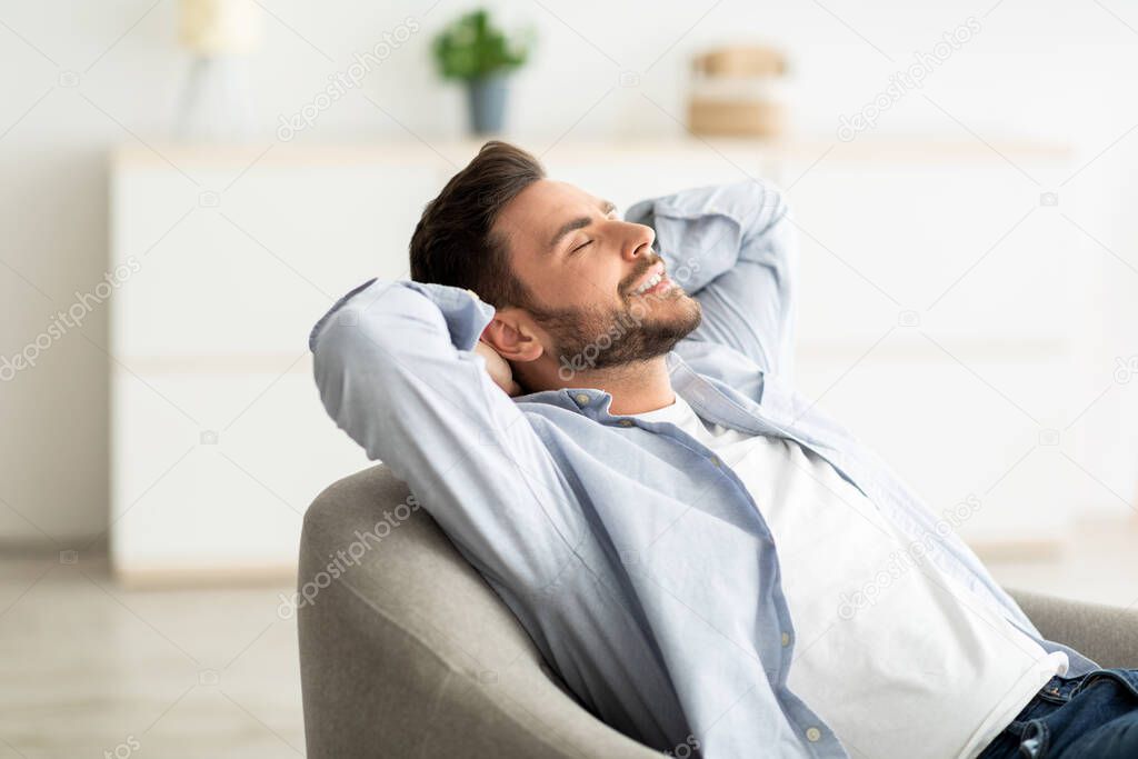 Lazy day. Happy relaxed man resting on comfortable armchair, holding hands behind head, sitting with closed eyes