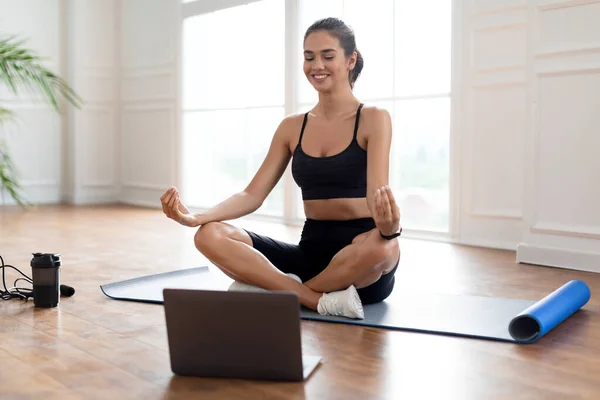 Young woman sitting on lotus position on mat with pc — Stock Photo, Image