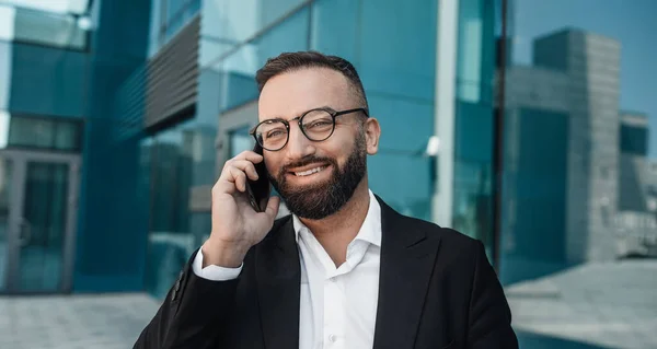 Діловий дзвінок. Впевнений бородатий бізнесмен спілкується на мобільному телефоні, стоячи на відкритому повітрі в місті — стокове фото
