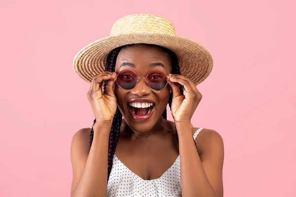 Excited millennial African American woman in summer wear touching her sunglasses, shouting WOW over pink background — Stock Photo, Image