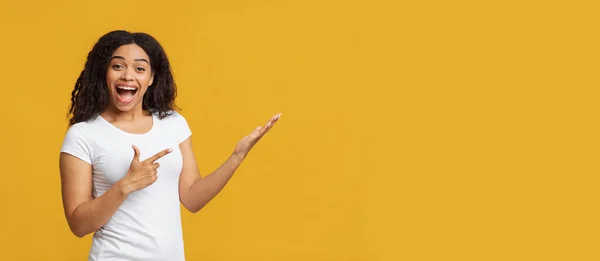 Kijk hier eens. Verheugd Afrikaans-Amerikaanse dame wijzend met vinger naar de vrije ruimte, staande op gele achtergrond, panorama — Stockfoto