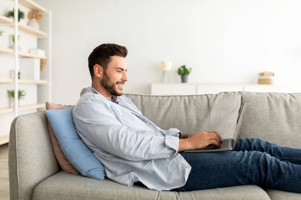 Glücklicher Millennial-Kerl mit Laptop-Computer, während er zu Hause auf dem Sofa ruht, Seitenansicht, freier Raum — Stockfoto