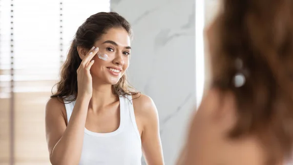 Mulher jovem aplicando creme facial hidratante pele lisa no banheiro — Fotografia de Stock