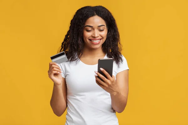 Fácil transferência de dinheiro. Sorrindo jovem afro-americana segurando cartão de crédito de débito e usando seu smartphone — Fotografia de Stock