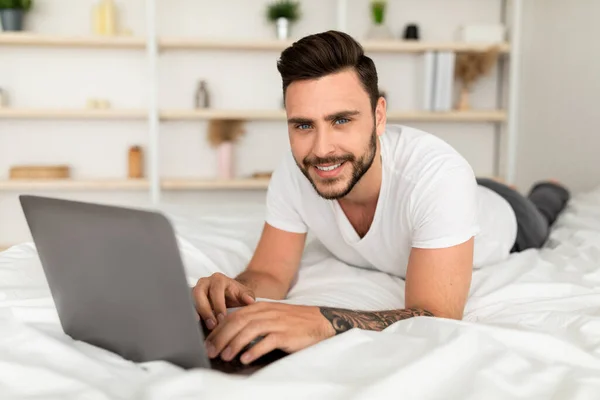 Praca w sypialni, przeglądanie w Internecie. Uśmiechnięty tysiącletni mężczyzna obudził się, pracując na laptopie, leżąc na białym łóżku w sypialni — Zdjęcie stockowe