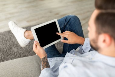 Genç adam boş siyah ekranlı dijital tablet kullanıyor, web sitesine göz atıyor ya da yeni uygulama gösteriyor, evdeki kanepede oturuyor