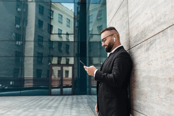 Empreendedor ouvir música em fones de ouvido sem fio e usando smartphone, de pé ao ar livre, espaço vazio, vista lateral — Fotografia de Stock