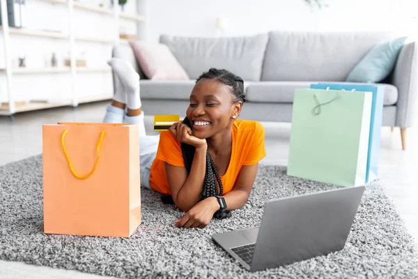 Mulher negra alegre com laptop pc e cartão de crédito deitado no chão, cercado por sacos de compras em casa — Fotografia de Stock