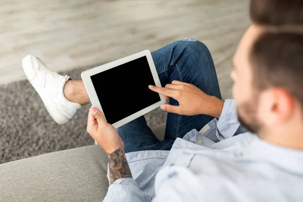 Joven usando tableta digital con pantalla en blanco y negro, navegando por el sitio web o mostrando una nueva aplicación, sentado en el sofá en casa — Foto de Stock