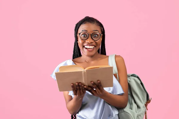 Excited young African American female student with backpack holding open book on pink background — стоковое фото