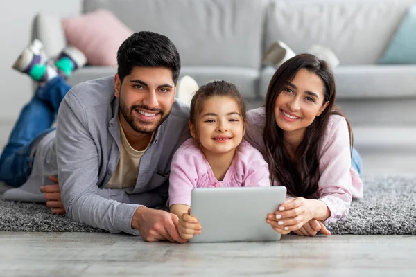 Giovani genitori arabi e la loro graziosa figlia utilizzando tablet digitale mentre sdraiati sul pavimento insieme e sorridenti alla fotocamera — Foto Stock