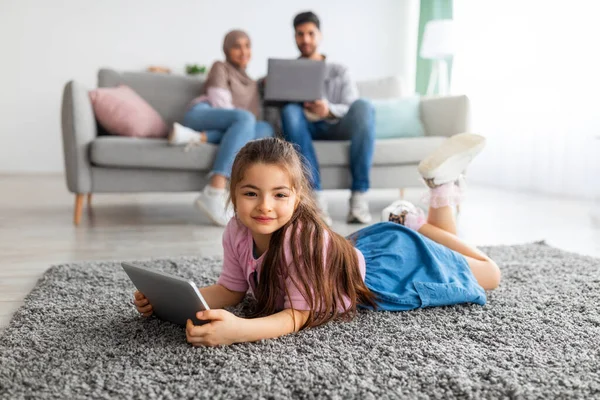 Leisure time. Cute little girl using digital tablet, lying on floor carpet and smiling at camera