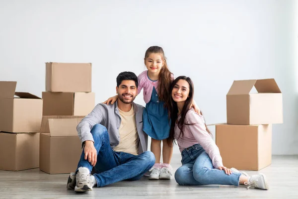 Весела східна сім'я з трьох чоловік переїжджає в новий будинок, сидячи серед картонних коробок у порожній вітальні — стокове фото