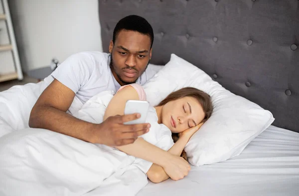 Celoso esposo negro revisando el teléfono inteligente de sus esposas, leyendo correspondencia mientras ella duerme en la cama en casa —  Fotos de Stock