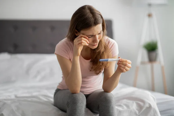 Olycklig vit kvinna tittar på graviditetstest, gråter på sängen hemma. Begreppet moderskapsproblem — Stockfoto