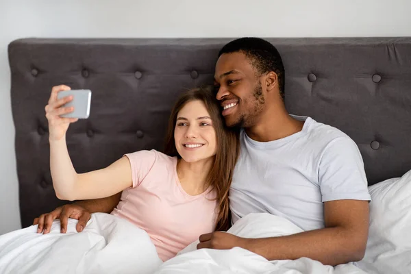 Millennial multiracial par med smartphone tar selfie i sängen innan du går och lägger dig, inomhus — Stockfoto