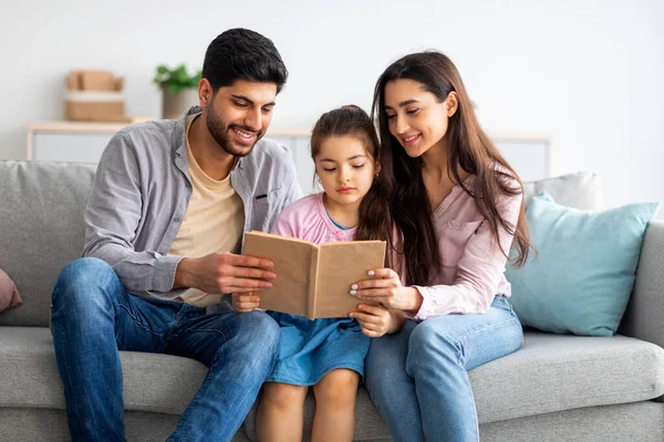 Familjefördriv. Glada arabiska föräldrar läser bok med sin lilla dotter samtidigt koppla av tillsammans på soffan — Stockfoto