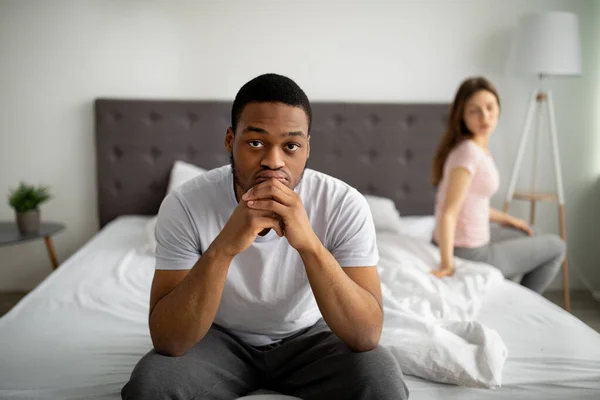 Szomorú fekete férfi ül az ágyon, mélyen elmerülve a gondolatokban, elárasztva családi problémákkal, elégedetlen barátnő a háttérben — Stock Fotó