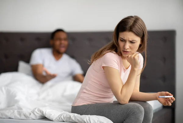 Jeune femme malheureuse assise sur le lit avec test de grossesse, mari noir lui criant dessus, à l'intérieur — Photo