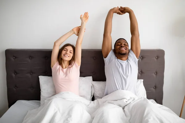 Νεαρό διαφυλετικό ζευγάρι που τεντώνεται στο κρεβάτι αφού ξυπνήσει το πρωί, σε εσωτερικούς χώρους — Φωτογραφία Αρχείου