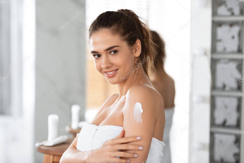Young Lady Applying Cream On Shoulder Moisturizing Skin In Bathroom