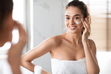 Yanaklarına krem süren genç bir kadın içeri yansımaya bakıyor.