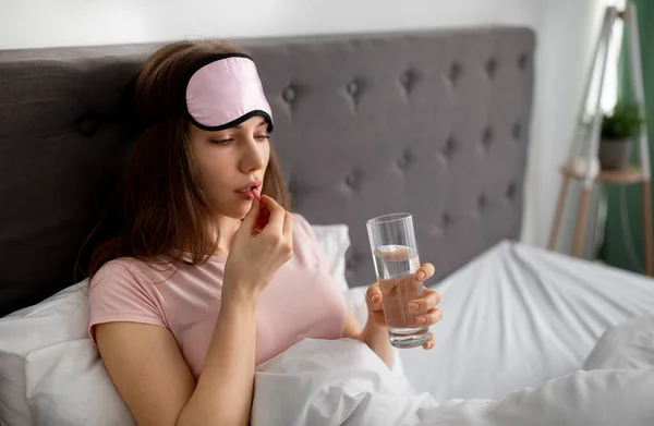Jovem sonolenta com copo de água tomando pílula na cama em casa, espaço de cópia — Fotografia de Stock