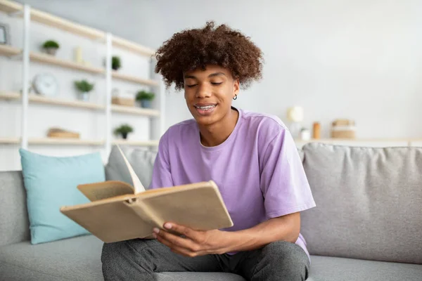 Adolescente positivo com livro aberto sentado no sofá confortável, estudando ou fazendo trabalhos de casa dentro de casa — Fotografia de Stock