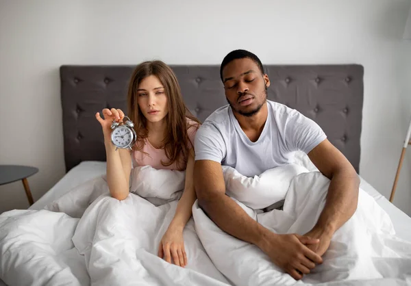 Ύπνο πολυφυλετικό ζευγάρι κάθεται στο κρεβάτι με ξυπνητήρι, δεν μπορεί να ανοίξει τα μάτια τους, πολύ κουρασμένος για να σηκωθεί το πρωί — Φωτογραφία Αρχείου