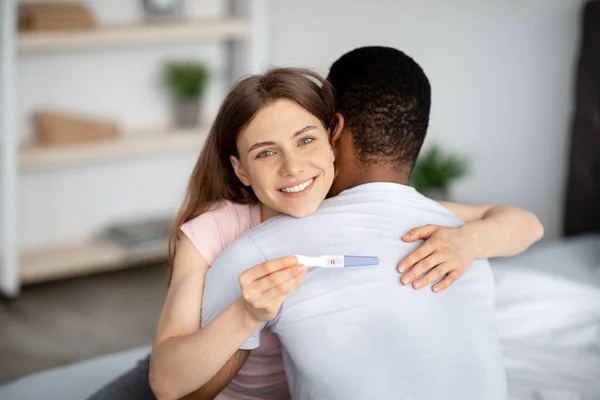 Verheiratetes multiethnisches Paar glücklich mit positivem Ergebnis des Schwangerschaftstests, umarmt einander zu Hause — Stockfoto