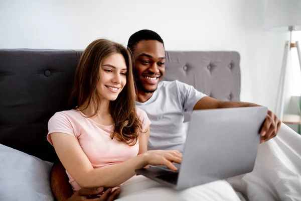 Glada unga multinationella par kopplar av i sängen, använder laptop, tittar på film tillsammans — Stockfoto