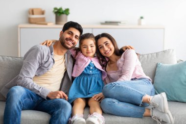 Aile evi eğlencesi. Mutlu doğu ebeveynleri ve tatlı kızları kanepede oturup kameraya gülümsüyorlar.