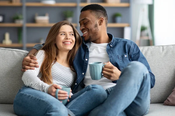 Feliz joven interracial pareja bebiendo café y abrazando en sofá en casa — Foto de Stock