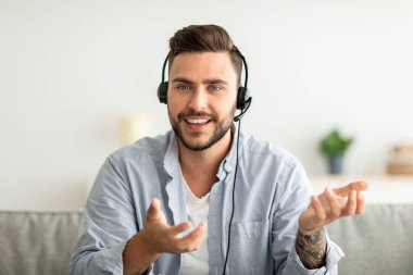 Kulaklıklı heyecanlı bir adam web kamerasıyla konuşuyor, uzaktaki meslektaşıyla video görüşmesi yapıyor, evdeki kanepede oturuyor..