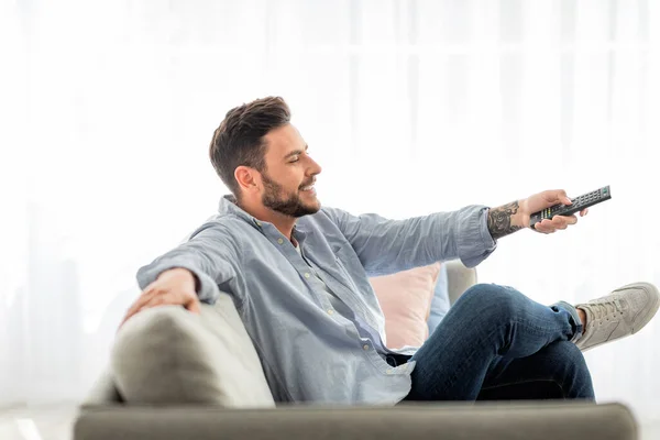 Relaxado homem alternando canais com controle remoto enquanto assiste tv em casa, relaxando no sofá, vista lateral — Fotografia de Stock
