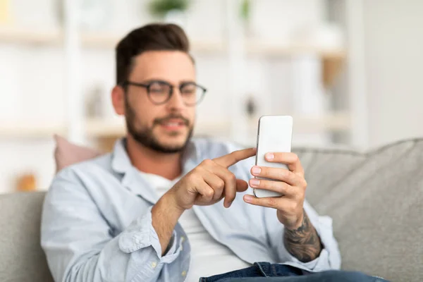 Красивий хлопець в окулярах використовує смартфон, торкаючись екрану, розслабляючись на дивані вдома, вільний простір — стокове фото