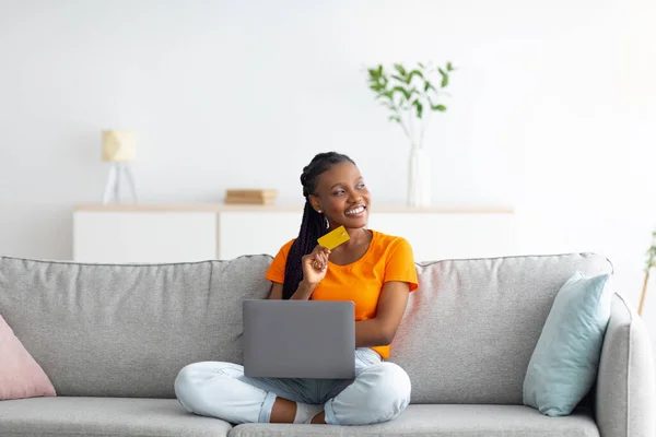 Giovane donna afroamericana che acquista online con carta di credito e laptop, seduta sul divano di casa, copia lo spazio — Foto Stock