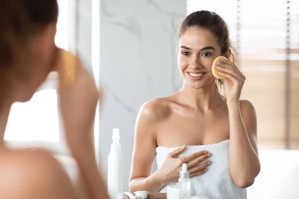 Mulher usando esponja cosmética desfrutando de rotina de cuidados com a pele facial no banheiro — Fotografia de Stock