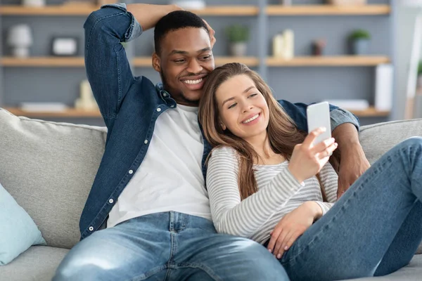 Portrét uvolněného mladého multiraciálního páru odpočívajícího s chytrým telefonem na gauči — Stock fotografie